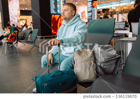 Young man sitting at airport gate waiting for a flight, looking out the window at airplanes with smartphone and suitcase. Modern air travel concept. Young man sitting at airport gate waiting for a flight, looking out the window at airplanes with smartphone and suitcase. Modern air travel concept. 136119870