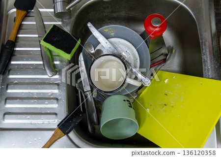 Cluttered kitchen sink with dirty dishes and sponges 136120358