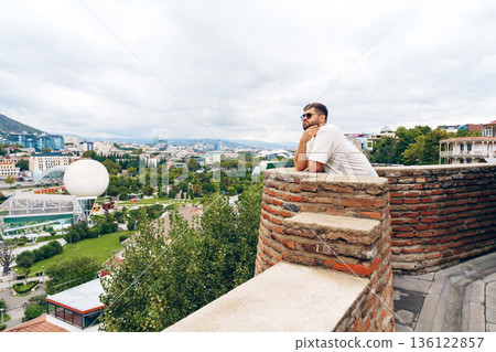Man looking over cityscape from wall in Tbilisi during cloudy day with greenery around 136122857