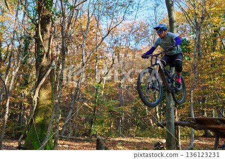 Mountain Biker Jumping Through Autumn Forest on Wooden Ramp During Trail Ride Adventure Mountain Biker Jumping Through Autumn Forest on Wooden Ramp During Trail Ride Adventure 136123231
