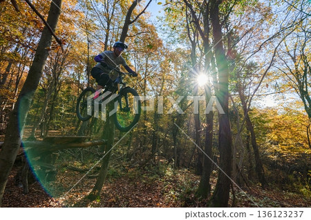 Mountain Biker Jumping From Wooden Platform in Autumn Forest With Sun Burst Mountain Biker Jumping From Wooden Platform in Autumn Forest With Sun Burst 136123237