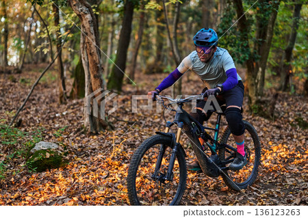 Mountain Biker Races Through Autumn Forest Trail in Protective Gear and Focused Expression 136123263