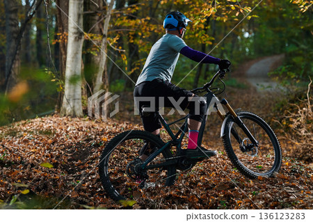 Mountain Biker on Forest Trail in Autumn Leaves Wearing Helmet and Protective Gear 136123283