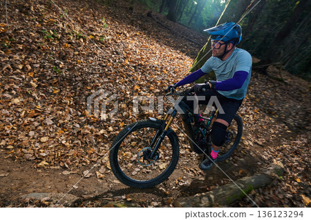 Mountain Biker Rides Through Leaf-Litter Trail in Autumn Forest With Gear, Helmet, and Knee Pads 136123294