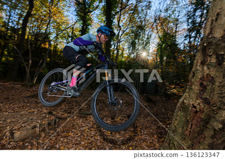 Mountain Biker Leaping Over Forest Trail in Autumn Sunlight and Falling Leaves 136123347