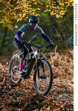 Mountain Biker Navigates Leafy Autumn Trail in Forest, Wearing Helmet and Protective Gear Mountain Biker Navigates Leafy Autumn Trail in Forest, Wearing Helmet and Protective Gear 136123530