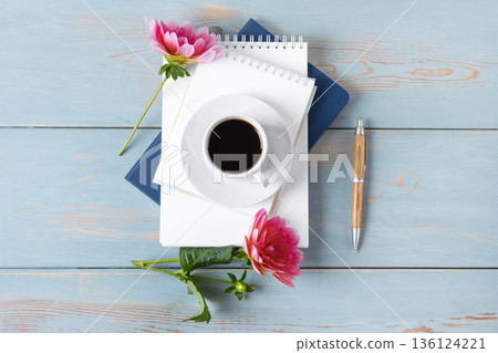 Top view of the desktop. Spiral notebook with an open blank page, pen, white cup with black coffee, dahlia flowers. Festive office desktop concept. Morning coffee cup. 136124221
