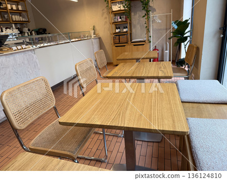 Modern cafe interior with wooden tables and woven chairs near window. Minimal design, natural materials, and cozy urban atmosphere Modern cafe interior with wooden tables and woven chairs near window. Minimal design, natural materials, and cozy urban atmosphere 136124810