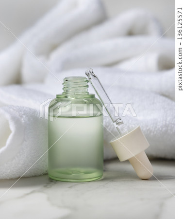 Pipette next to opened green glass dropper bottle near white bath towel, cosmetic mockup 136125571