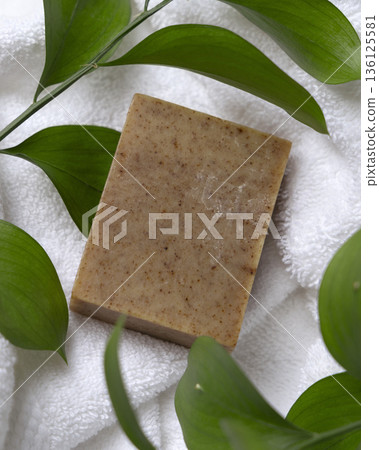 Unbranded brown soap bar on white terry towel with green leaves, cosmetic packaging mockup 136125581