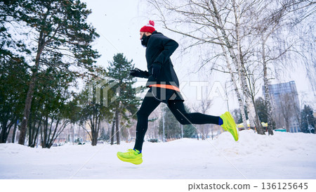 Man running in snowy city park wearing winter sportswear. Man running in snowy city park wearing winter sportswear. 136125645