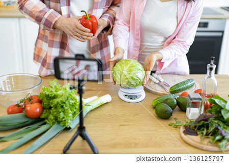 A couple prepares a cooking video with fresh vegetables on a wooden table in a well-lit kitchen A couple prepares a cooking video with fresh vegetables on a wooden table in a well-lit kitchen 136125797