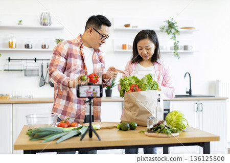A couple is filming a cooking vlog in their kitchen, showcasing fresh vegetables and healthy eating habits 136125800