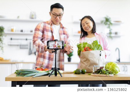 An Asian couple records a cooking video in their bright, modern kitchen, showcasing fresh ingredients An Asian couple records a cooking video in their bright, modern kitchen, showcasing fresh ingredients 136125802