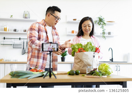 A couple is filming a cooking vlog in their kitchen, showcasing fresh vegetables from a grocery bag 136125805