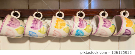 Ceramic mugs with floral motifs hanging on kitchen rack 136125935