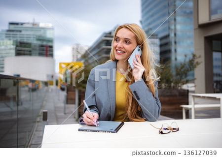 Businesswoman talking on phone and writing on tablet outdoors 136127039