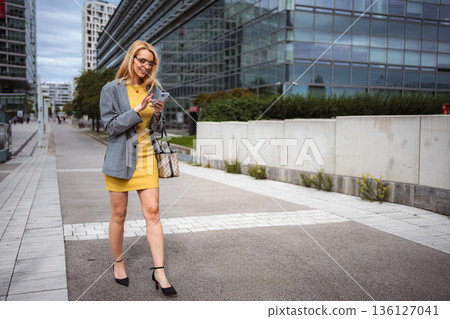 Businesswoman walking in city while using smartphone 136127041