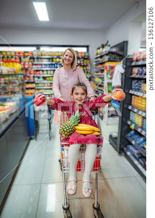 Mother pushing daughter in supermarket shopping cart with fresh fruit 136127106