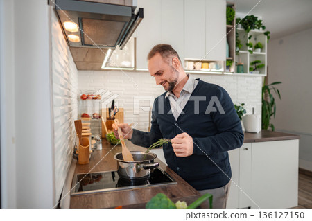 Happy man cooking meal adding fresh herbs in kitchen 136127150