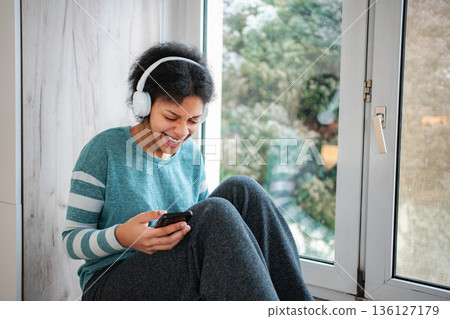 Young woman enjoying music streaming on smartphone near window 136127179
