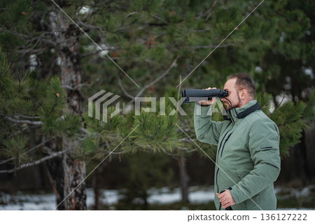 Man birdwatching with binoculars in pine forest during winter Man birdwatching with binoculars in pine forest during winter 136127222