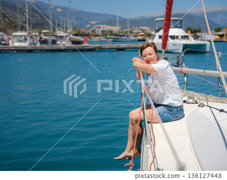 Mature woman relaxes on private yacht during sunny day in Finike Marina. Turkish coastline and luxury boats surround sailing vessel in Mediterranean port during summer vacation. Mature woman relaxes on private yacht during sunny day in Finike Marina. Turkish coastline and luxury boats surround sailing vessel in Mediterranean port during summer vacation. 136127448