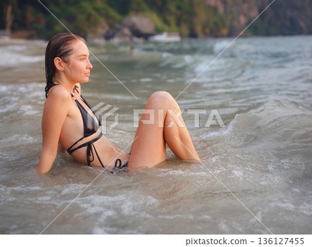 Young woman enjoying serene evening at sea in Krabi, Thailand during sunset. Calm waves, golden light, and peaceful atmosphere highlight tropical beauty and connection with nature. Young woman enjoying serene evening at sea in Krabi, Thailand during sunset. Calm waves, golden light, and peaceful atmosphere highlight tropical beauty and connection with nature. 136127455