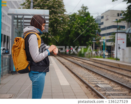 Tourist woman stands at tram stop in Frankfurt, waiting for public transportation amidst urban setting, surrounded by city life and modern buildings, experiencing everyday rhythm of city. Tourist woman stands at tram stop in Frankfurt, waiting for public transportation amidst urban setting, surrounded by city life and modern buildings, experiencing everyday rhythm of city. 136127529