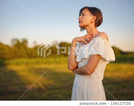 Beautiful woman in white dress standing in summer field during golden sunset in Croatia, warm sunlight, romantic evening mood, natural countryside atmosphere, soft wind and peaceful scene Beautiful woman in white dress standing in summer field during golden sunset in Croatia, warm sunlight, romantic evening mood, natural countryside atmosphere, soft wind and peaceful scene 136127530