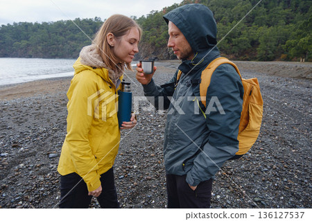 Couple enjoys Turkish coastline. Sitting on stones, watching sea waves, relaxing together, slow romantic moment near calm blue water under soft winter light. 136127537