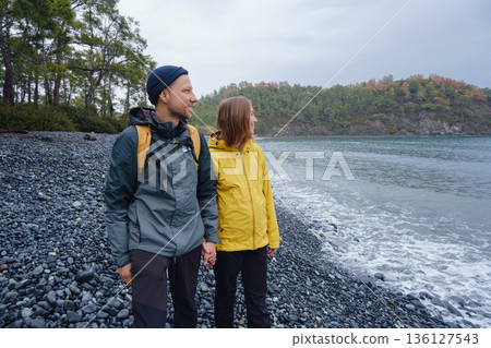 Couple enjoys Turkish coastline. Sitting on stones, watching sea waves, relaxing together, slow romantic moment near calm blue water under soft winter light. Couple enjoys Turkish coastline. Sitting on stones, watching sea waves, relaxing together, slow romantic moment near calm blue water under soft winter light. 136127543