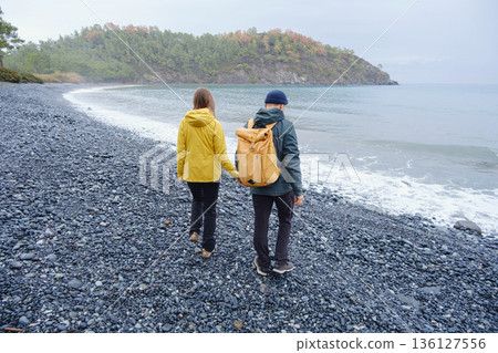 Couple enjoys Turkish coastline. Sitting on stones, watching sea waves, relaxing together, slow romantic moment near calm blue water under soft winter light. Couple enjoys Turkish coastline. Sitting on stones, watching sea waves, relaxing together, slow romantic moment near calm blue water under soft winter light. 136127556