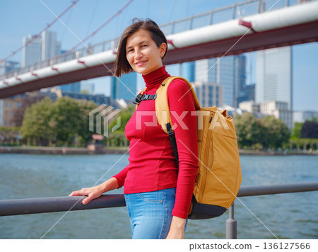Tourist woman explores modern Frankfurt, walking through vibrant streets with contemporary architecture, skyscrapers, and urban spaces, enjoying lively atmosphere of modern side. 136127566