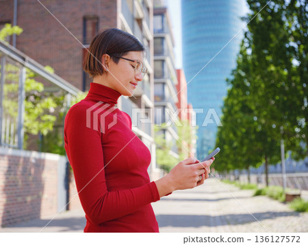 Beautiful woman in walking in Frankfurt on spring day and holding mobile phone. Urban lifestyle concept. Check social networks, send sms or book hotel, exuding confidence and professionalism 136127572