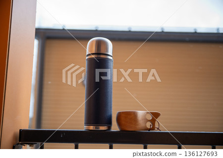 Stainless steel thermos bottle and wooden cup on balcony in warm morning light Stainless steel thermos bottle and wooden cup on balcony in warm morning light 136127693