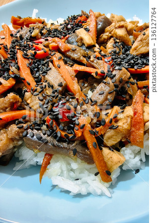 Chicken stir fry is prepared with carrots, mushrooms, and black sesame seeds, served on a plate of white rice for a tasty meal enjoyed at home 136127764