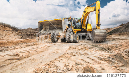 Construction equipment, an excavator and a loader, are actively working on a construction site, moving earth embankments in a large open area under a partially cloudy sky Construction equipment, an excavator and a loader, are actively working on a construction site, moving earth embankments in a large open area under a partially cloudy sky 136127773