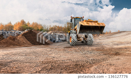 Loader operates at a construction site, moving dirt and rocks around. The sky is cloudy and autumn trees are in view. Work continues on the site Loader operates at a construction site, moving dirt and rocks around. The sky is cloudy and autumn trees are in view. Work continues on the site 136127776