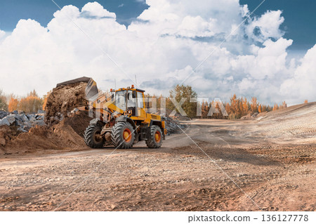 A loader is lifting and moving dirt at a construction site. The ground is uneven with rocks and material. Trees are visible in the background under a cloudy sky 136127778