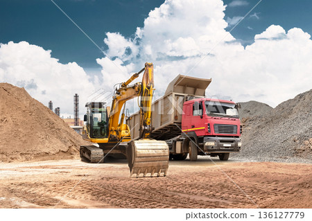 An excavator moves sand from a mound into a truck at a busy construction site. Heavy equipment performs earthmoving and bulk material handling under a clear blue sky 136127779