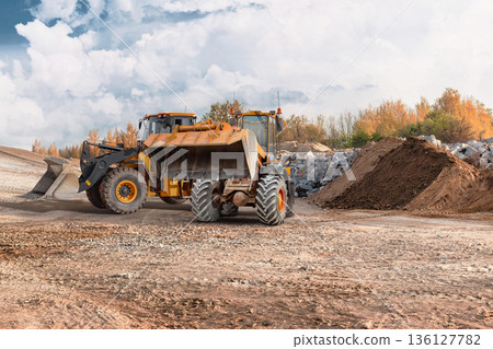 Loader lifts and moves dirt while excavator works on the site. Both machines are used for construction tasks. The scene is active with earth moving Loader lifts and moves dirt while excavator works on the site. Both machines are used for construction tasks. The scene is active with earth moving 136127782