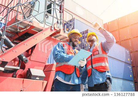 Two African American logistic workers wearing reflective vests and white helmets talk about logistics operations at shipping container yard. Transportation import and export logistic industry concept. 136127788