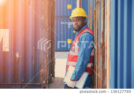 African American logistic workers wear yellow helmets and reflection shirts, hold document paper and look to check inventory or job details at the shipping container yard. Transportation import and African American logistic workers wear yellow helmets and reflection shirts, hold document paper and look to check inventory or job details at the shipping container yard. Transportation import and 136127789