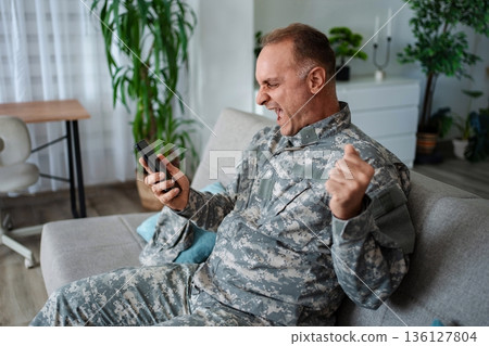 Excited military man receiving good news on smartphone at home 136127804