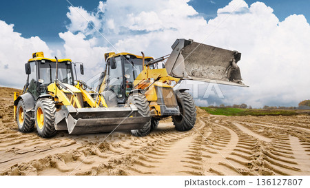 Two construction loaders are ready to go to work at a construction site, and a large pile of sand is visible against the cloudy sky. The work continues Two construction loaders are ready to go to work at a construction site, and a large pile of sand is visible against the cloudy sky. The work continues 136127807