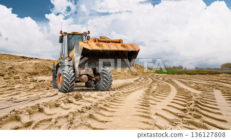 A loader or bulldozer with its bucket raised stands ready to work, while in the background a sand pile is visible, indicating construction work underway under a cloudy sky A loader or bulldozer with its bucket raised stands ready to work, while in the background a sand pile is visible, indicating construction work underway under a cloudy sky 136127808