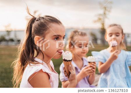 Three little girls enjoying ice cream cones in a park during sunset 136127810