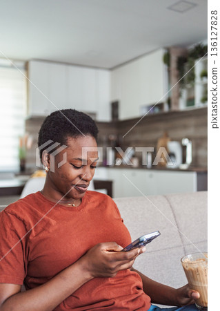 Young black woman using mobile phone while drinking coffee at home 136127828