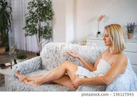 Woman applying moisturizing body lotion after shower, sitting on sofa 136127843